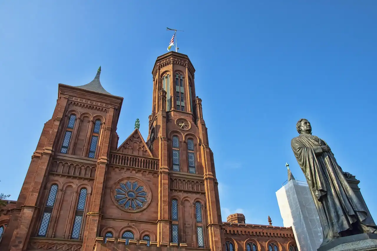 The red-brick towers of the Smithsonian Castle on the National Mall under a blue sky, with a statue beside it.