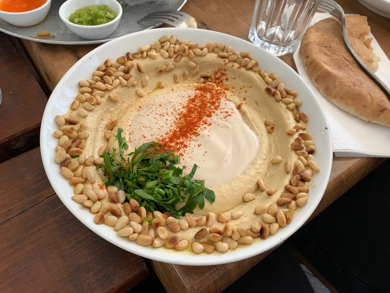 A plate of hummus topped with pine nuts, paprika and herbs, with warm pita on the side