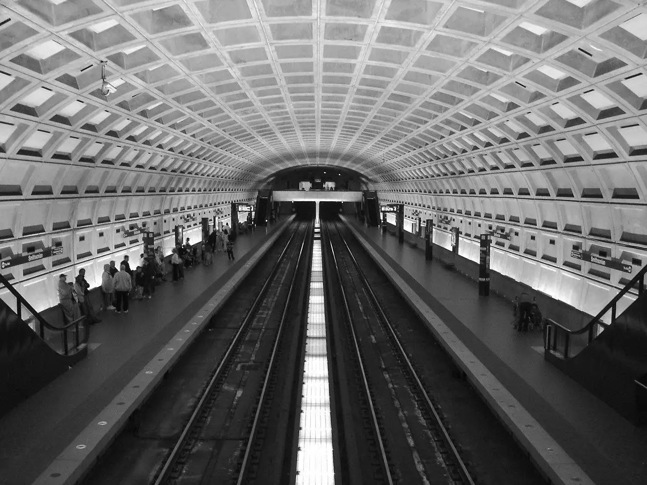 A black-and-white view down the platform of a Washington Metro station, under its vaulted concrete ceiling of coffered squares.