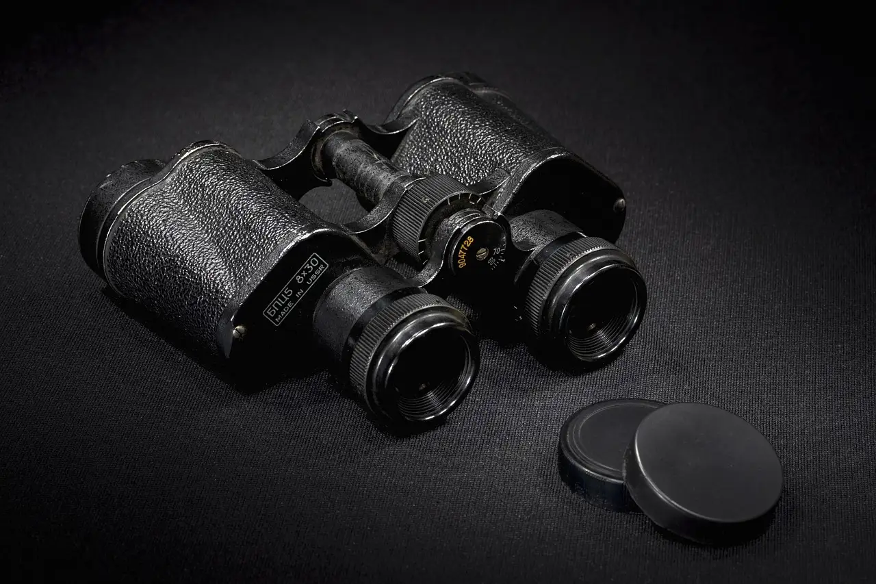 A pair of old black binoculars with their lens caps beside them on a dark surface.