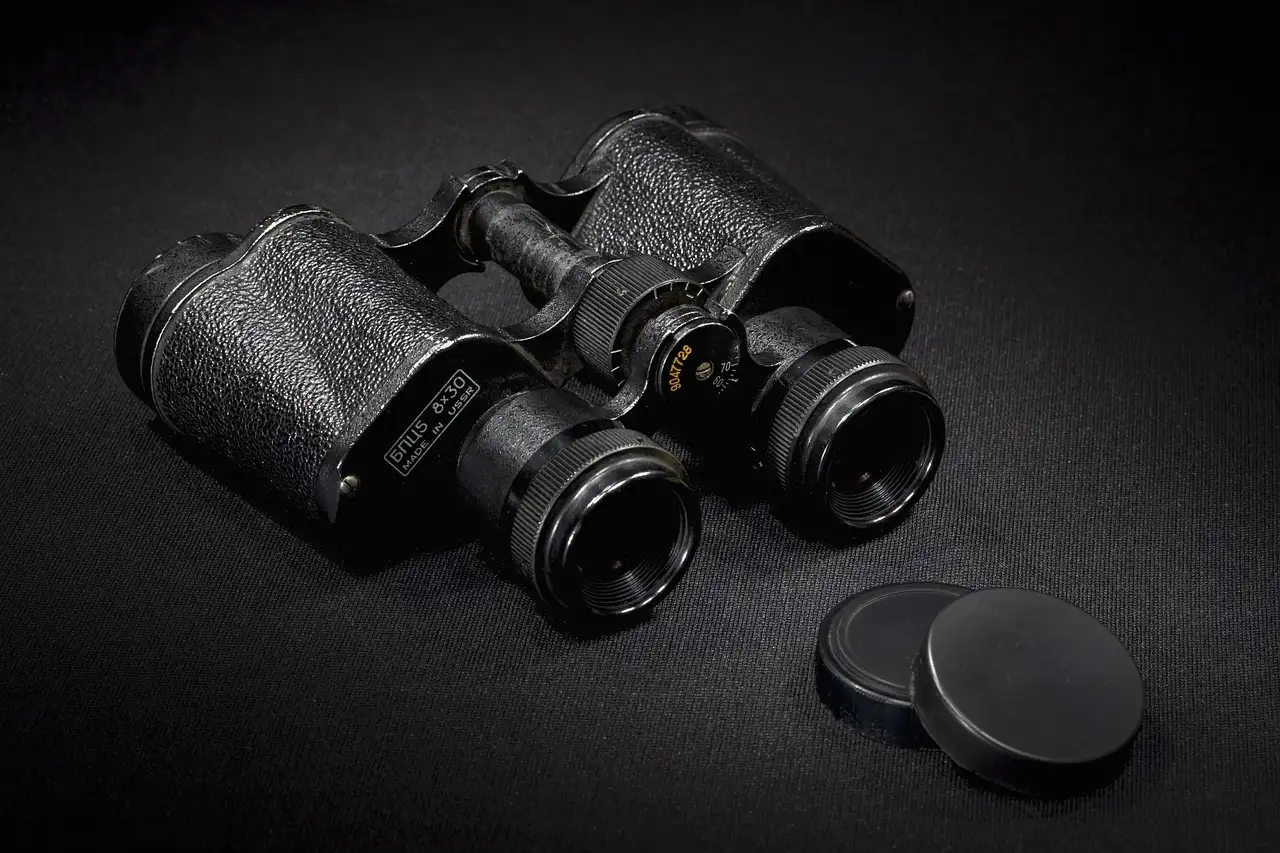 A pair of old black binoculars with their lens caps beside them on a dark surface.