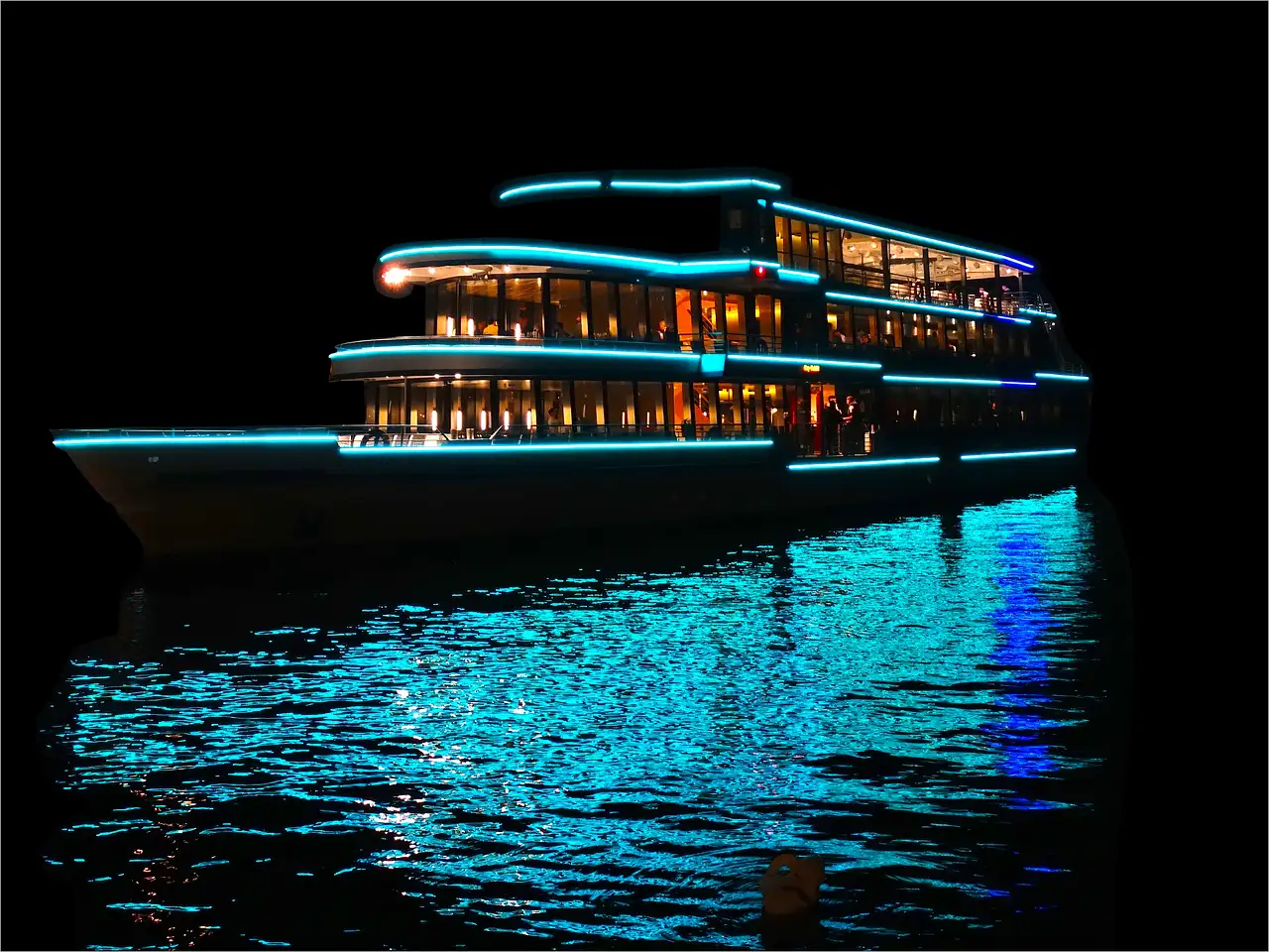 A glass-sided tour boat lit up at night, its lights reflected in the dark water