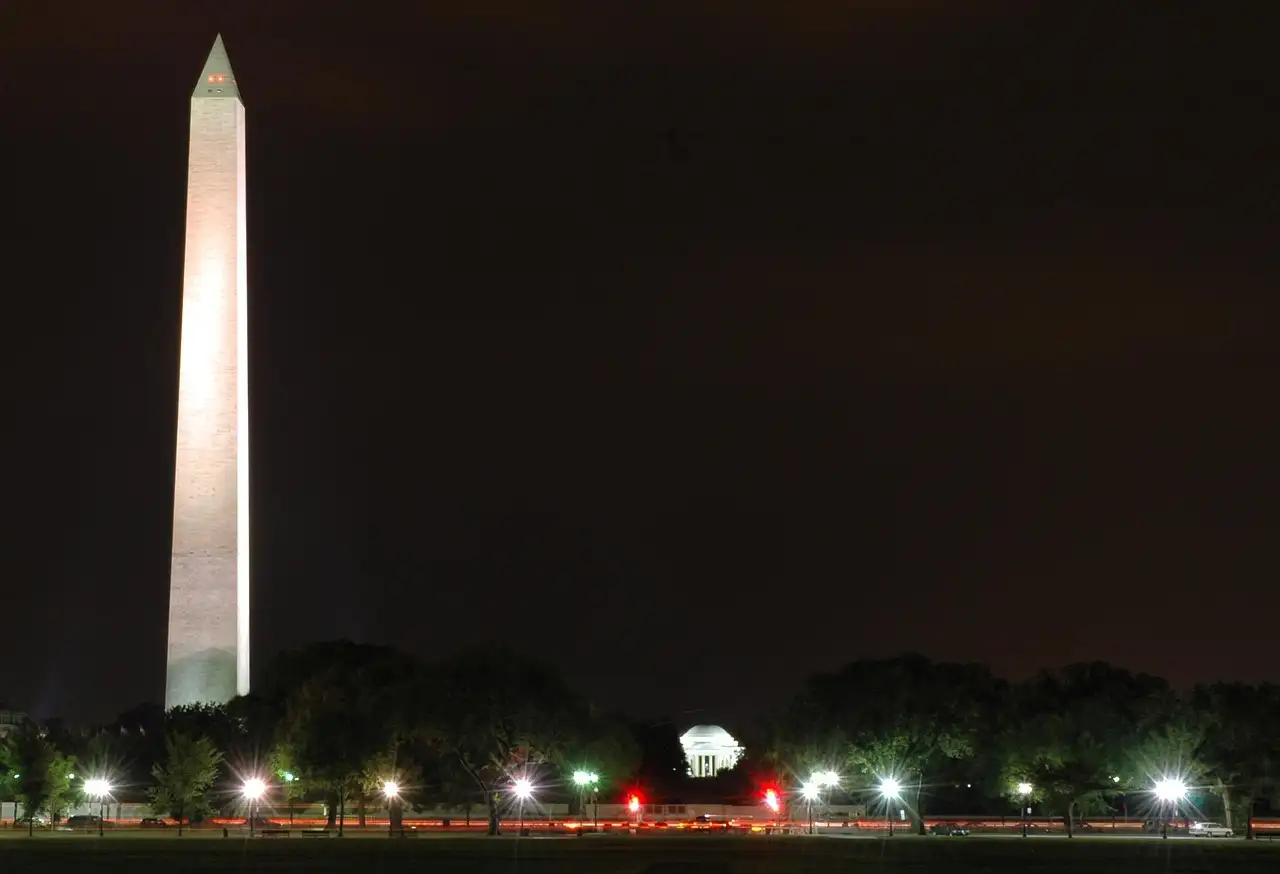The Washington Monument lit white against a dark night sky, with a row of lights below