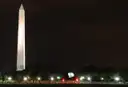The Washington Monument lit white against a dark night sky, with a row of lights below